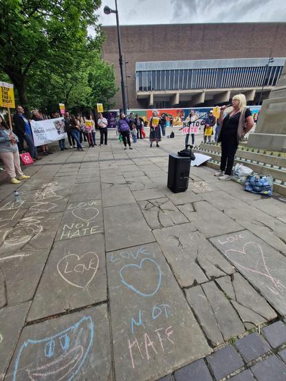 A closer look at the paved area and murals described in the previous image. There are lots or hearts drawn in chalk. The nearest paving slab to the camera reads "LOVE NOT HATE". The sound system and mic and still visible in the background, as is part of the cenotaph. A white lady with blond hair is delivering a speech.