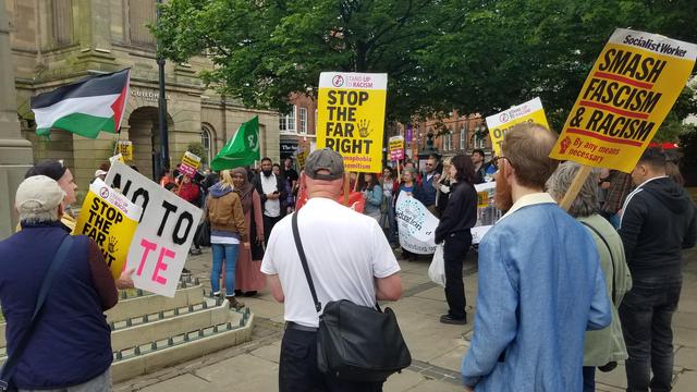 A view from the crowd shows people from behind holding signs including "SMASH FASCISM AND RACISM", "STOP THE FAR RIGHT", and "NO TO HATE".
