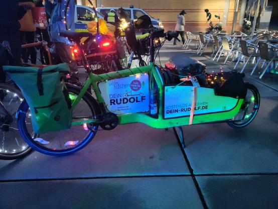 Ein grünes Bullitt ist mit bunten Lichterketten beleuchtet. Im Hintergrund stehen weitere bunt beleuchtete Fahrräder