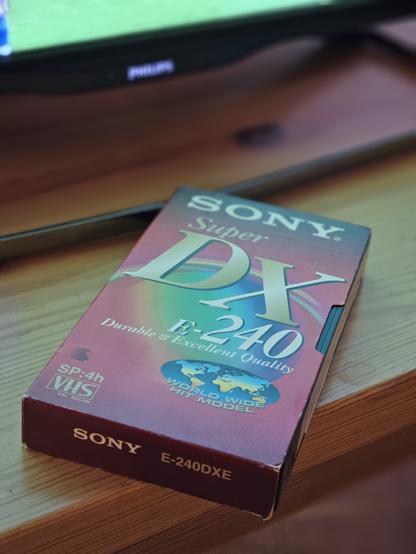 Rote Sony-Videokassette mit 240 Minuten Laufzeit vor dem Fernseher auf einem Holztisch.