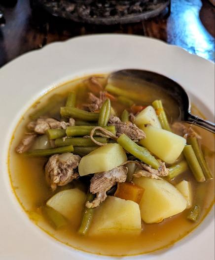 Weißer Suppenteller auf dunklem Holztisch. Im Teller eine klare, rötlich-braune Fleischbrühe. Die Einlage besteht aus Kartoffelstücken, grünen Bohnen, Fleisch und Tomatenstücken. Ein silberner Suppenlöffel liegt im Teller.