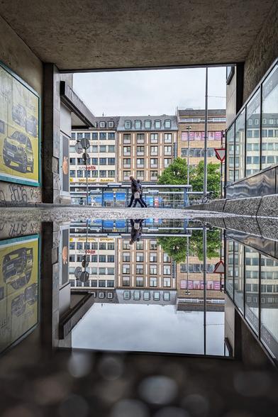 Das Bild zeigt eine städtische Szene, die durch eine Pfütze auf dem Boden gespiegelt wird. Im Vordergrund gibt es einen überdachten Bereich, der einem Tunnel ähnelt. Links im Bild ist ein Werbeplakat für ein Auto zu sehen. Zwei Menschen gehen in der Mitte des Bildes und ihre Silhouetten spiegeln sich in der Pfütze. Im Hintergrund erstrecken sich mehrstöckige Gebäude mit zahlreichen Fenstern und Geschäften im Erdgeschoss. Die Spiegelung schafft eine fast symmetrische und visuell spannende Komposition, die das Bild besonders macht.