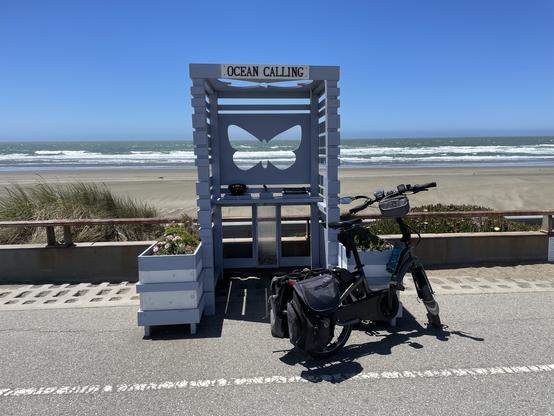 A bicycle is parked in front of a phone-booth-sized structure along the side of the roadway of the former Great Highway in San Francisco, now Sunset Dunes Park. A sign at the top says Ocean Calling. There is a cutout of a butterfly on the back wall. It’s open in the front, and has open slats on the side. Planters help weigh it down on either side of it. There is a shelf inside. A close-up photo of that shelf follows this one. 

Behind the phone booth are some dunes, a wide almost flat sandy beach, some rolling waves, and a big blue beautiful sky. Not seen: it’s windy!