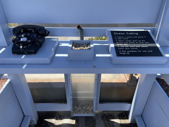 Inside the phone booth on the shelf is an old rotary telephone, and a small box of pebbles with a pipe dropping vertically behind it to a plexiglass box beneath it. There are many pebbles in the box. On the right side is a plaque that says:
Ocean Calling
1. Select a stone and place it in the chute.
2. When ready, pick up the phone and speak to your lost loved one.
3. Let the wind and waves carry your words. 
4. Say goodbye for now and hang up.