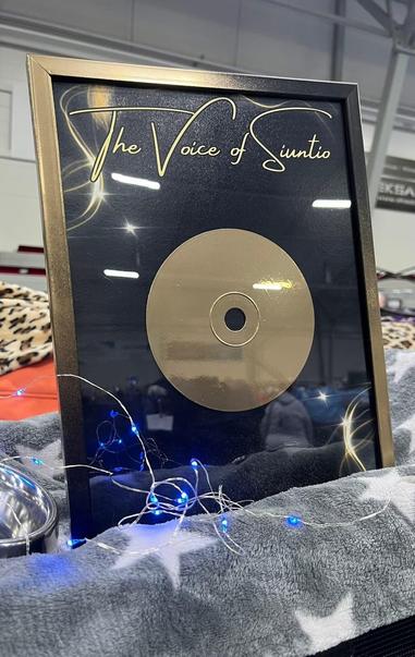A framed award plaque featuring a gold disc labeled "The Voice of Siuntio." The plaque is placed on a soft, star-patterned blanket, with decorative blue string lights nearby and a small bowl visible.