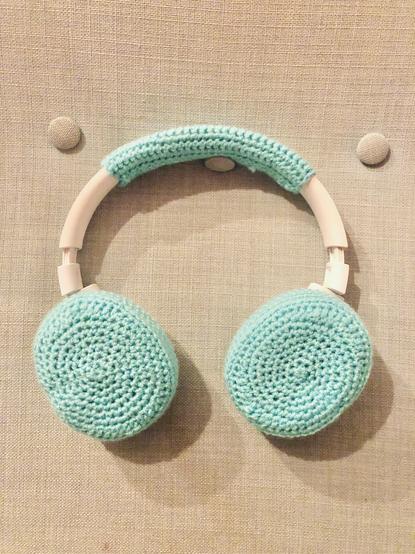 White wireless over the ear headphones with crocheted turquoise covers over the ear pieces and the soft bit at the underside of the arch. The headphones are hanging on a button of a light turquoise chair, but the rest of the chair isn’t visible. The photo is quite poorly lit, since it was taken inside around 10pm.