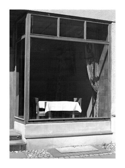 The black and white photo shows a store with a large display window. Inside is a table with two chairs, covered with a white tablecloth. Part of the window is covered by a curtain. The overall impression is quiet and somewhat deserted.