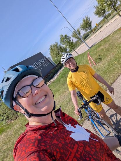 Two cyclists in front of an industrial park sign (says something like Anthony Henday Business Parki think) with green grass, some trees, and a beautiful blue sky in the background.  Cyclist in the foreground is wearing a red cycling jersey with a white maple leaf, teal helmet, and tan mirrored sunglasses.  Cyclist in the background is wearing a yellow Star Trek The Original Series cycling kit, with a white helmet, holding a blue road bike.