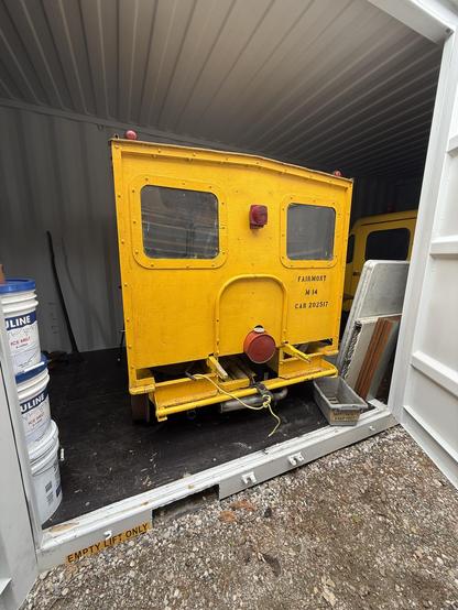A Fairmont M14 speeder, a boxy looking bright yellow rail vehicle that could hold around 4 riders, sitting inside a shipping container with the rear facing an open door