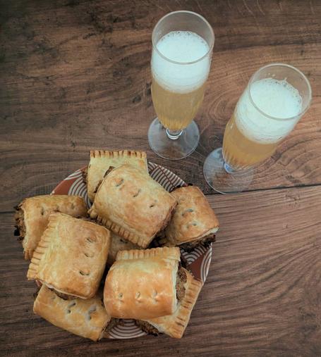 A photo from above of a plate of vegetarian sausage rolls, with two champagne flutes containing Bellini cocktails next to it, all sitting on a brown wooden background. The sausage rolls have golden pastry with three little cuts across the top and a very meaty looking brown filling. The Bellinis are light-peach coloured with white foam top.