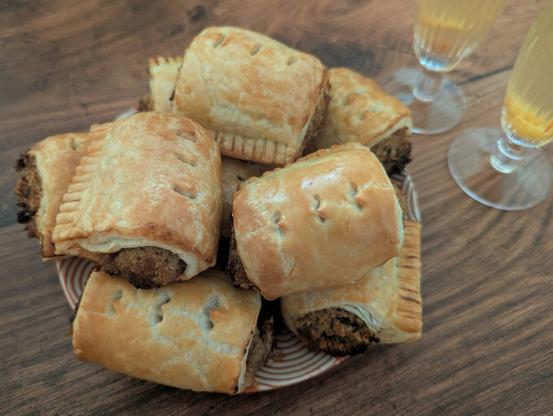 A close-up photo of a plate of vegetarian sausage rolls, with the bottom edge of some champagne flutes containing Bellinis in the top corner, all sitting on a brown wooden background. The sausage rolls have golden pastry with three little cuts across the top and a very meaty looking brown filling. The Bellinis are light-peach coloured with some peach pulp at the bottom.