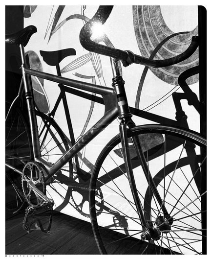 Schwarzweißfotografie: ein silbernes Rennrad aus den 1980er Jahren (DDR Wettkampfrad von Textima / Diamant) steht in einem Schaufenster, durch Sonneneinstrahlung wirft diese hinter das Fahrrad noch einmal einen verzerrten Schatten des Rades, außerdem einen hellen Fleck der Spiegelung auf die Schaufensterscheibe