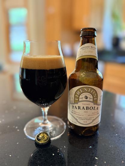 A tall snifter glass full of dark beer with a tan head. The brown bottle is to the right with a tan neck label and a tan body label with gold accents and black and white lettering.