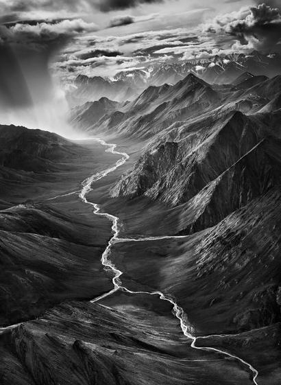 Das Foto zeigt die Brooks Range, Alaska, aufgenommen von Sebastião Salgado. Ein dramatisches Foto mit einem Wolkenbruch auf der linken Bildseite, schroffem Gebirge im Hintergrund und einem Fluss, der sich vom Auge des Betrachters in Richtung des Wolkenbruchs entlang zieht.
