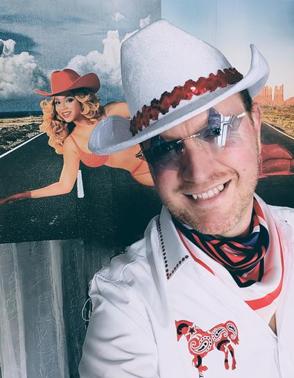 My outfit for Cowboy Carter: white cowboy hat with red trim, white shirt with a red paisley horse on the side, a red, white & blue neckerchief & blue star-shaped glasses; a Beyoncé poster behind me