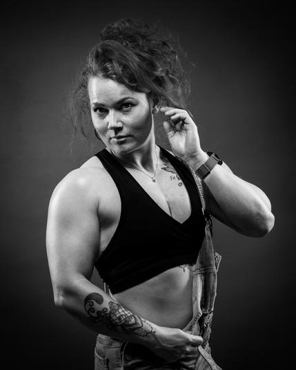 The image is a black and white portrait of a person posing with one hand near their face. They have a muscular build, wear a sleeveless top, and have tattoos on their arm. Their hair is styled up and they appear to be wearing overalls.