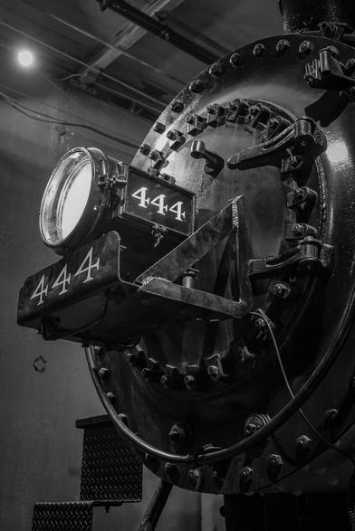 The image is a black and white photograph of a steam locomotive's front section, specifically focusing on the headlamp and the number plate. The headlamp is circular with a clear glass cover, mounted on a metal bracket. The number "444" is prominently displayed in white on a black background, both on the headlamp's plate and on a separate metal plate below it. The headlamp and number plate are secured with numerous bolts, and the metal surface shows signs of wear and rust, indicating age and use. The background is dimly lit, with a single light source visible on the ceiling, casting a soft glow on the scene. The overall composition emphasizes the industrial and historical significance of the locomotive.

Provided by @altbot, generated privately and locally using Ovis2-8B