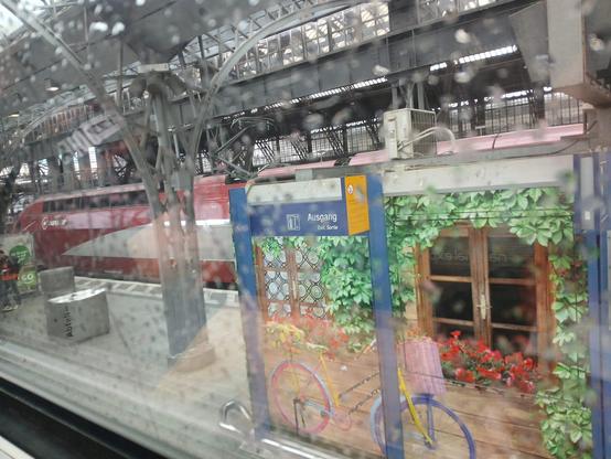 Blick am Bahnhof aus einem Zugfenster mit Regentropfen auf der Scheibe. Zu sehen ist der Bahnhof mit Metall-Glas-Dach, ein roter Eurostarzug am Nachbargleis und eine Art Hütte auf dem Bahnsteig mit aufgeklebtem bunten Fahrrad-Fenster-Blumen-Efeu-Bild.
