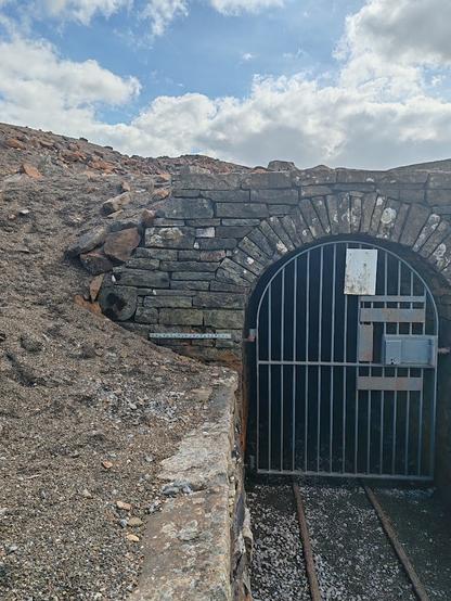 The entrance to Carrs Mine, Nenthead. There is a metal bar gate across a barrel vaulted dry stone entrance. Iron rails still in place on the approach as if wagons could run in and out.