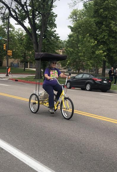 A smiling person riding a yellow trike with a canopy down a quiet street. She is wearing a purple shirt with a bright green and pink print.