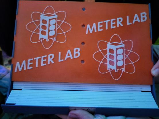 Ein Spender für Meterstäbe wird vor die Kamera gehalten. Er hat das Metalab-Logo und dazu den Text "Meterlab" zwei Mal darauf. Der Spender ist mit mehreren Metern bestückt.