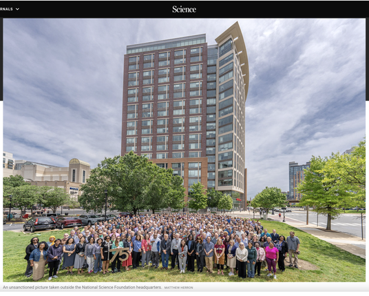 Photo to mark the 75th anniversary of the US National Science Foundation, with 250 staffers posed to the side of the building in a non-official photo taken on the staff time by an amateur photographer so that the senior management who were not allowed to be present could allow this short event.  The building is about 18 stories tall, the meeting area is all bright green grass and there are other buildings and a cluttered landscape around the people and building, with cloudy skies.