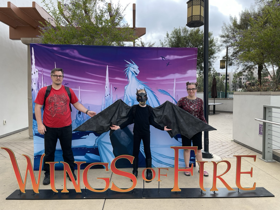A white man wearing a red shirt, a kid dressed as a black dragon, and a white woman standing in front of a picture of a white dragon, with the words “Wings of Fire” in front
