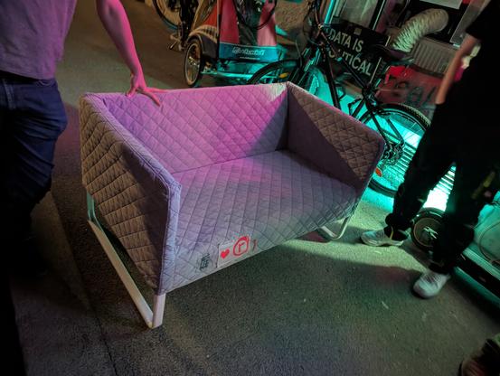 Ein kleines Sofa steht auf dem Gehsteig. Es ist grau und an der Vorderseite der Sitzfläche ist das Metalab-Logo, ein Herz und dann das Logo des realraums.