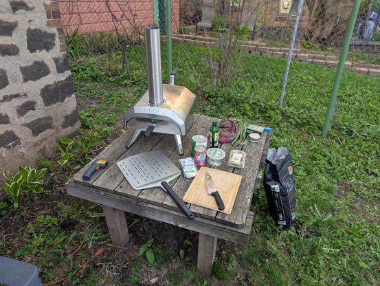Wooden table with a portable pizza oven and all of the necessary ingredients for a pizza.