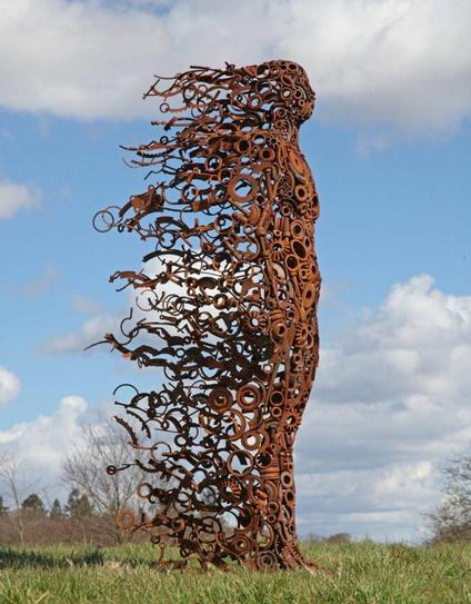 A tall sculpture of a woman made from rusty cogs and metal pieces set in a field. Photographed side on the impression of her body being blown in a windswept manner