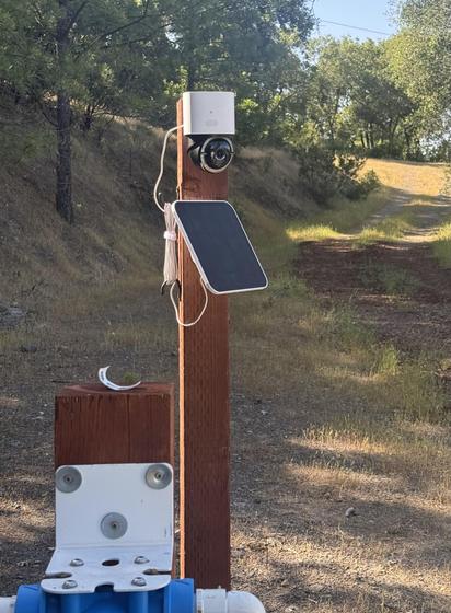 A pan & tilt camera and solar panel attached to a pressure treated 4”x4” post. Trees and a dirt road are in the background
