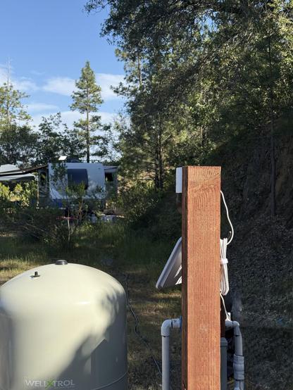 Back view of the camera post. A white pressure tank is in the lower left of the frame. Beyond the post are trees and our RV