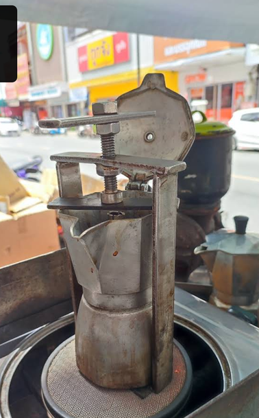 Moka coffee pot located on a street coffee vendor cart.
It has some kind of custim metal clamp arrangement holding the top bit on.
There are Thai shops and city streetscape in the background.