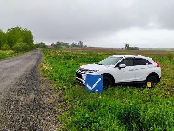 A white Mitsubishi SUV sits in a field at the side of a gravel road. Acting as a rally checkpoint, there is a blue and white CO board at the front of the vehicle. It's wet and the skies are grey and foreboding, a but the fields are green with the promise of a good growing season.