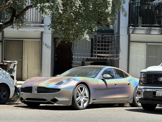An iridescent car is parked on the street in front of an apartment building. The vehicle features striking colors that shift in the light. Nearby, a white car and a black pickup truck are visible. In the background, a tree provides some shade