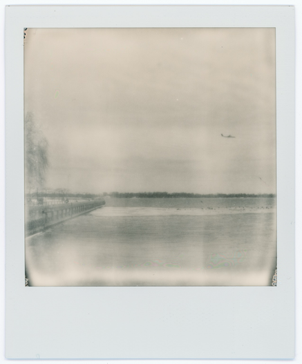 A fading black and white Polaroid depicting a former industrial wharf converted to urban beach park as a plane flies low overhead. A strip of ice is a landing zone for waterfowl.