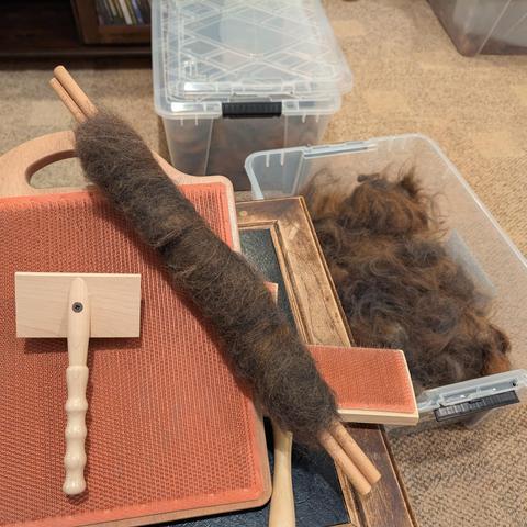 A rollag of sorts of dark fur with brownish hints wrapped around two dowels into a long sausage like shape. It sits on a fiber blending board, with a bin of dark gray and browinsh fur off to the side. The fiber has just been combined on the blending board and then rolled up around the dowels.