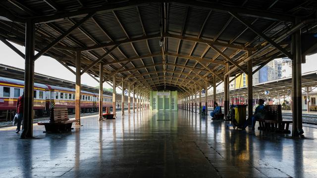 A metal roof covered passenger platform with empty rail car on both sides. a line of sitting benches with some train station workers sitting on them.