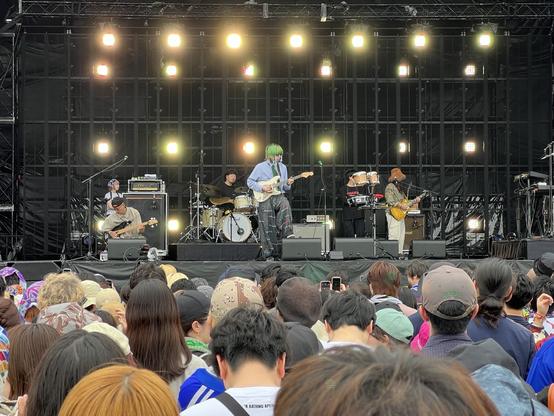 A live music performance featuring Kroi on stage with a guitarist in the foreground, surrounded by musicians playing various instruments. The audience is visible in the foreground, engaging with the performance. Bright stage lights illuminate the scene.