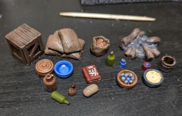A bunch of little boxes, bottles, food, campfire, and a book, all miniature scale. The book has the Delicious in Dungeon dragon painted on it