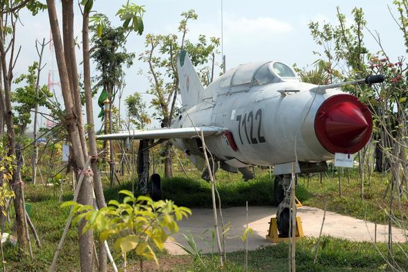Photo shows a fighter jet placed on a concrete platform and surrounded by green trees and vegetation, slowly growing and envelop the weapons of war. It is a sunny day, and the sky is lightly clouded and light blue.