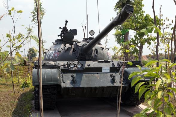 Photo shows an old main battle tank placed on a concrete platform and surrounded by green trees and vegetation, slowly growing and envelop the weapons of war. It is a sunny day, and the sky is lightly clouded and light blue.