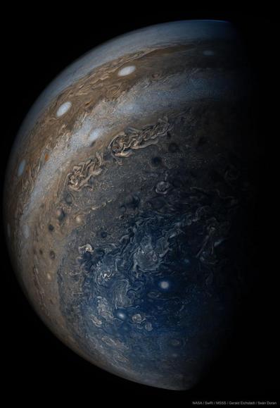 A close-up image is shown of the planet Jupiter. Many clouds are visible including clouds colored blue near the bottom, on the left, and white oval clouds on the upper right.