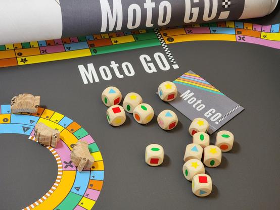 A brightly-coloured race track game with hand-carved wooden motorcycle game pieces, wooden dice with shapes instead of numbers, a rulebook, and a packaging tube. Many components feature the name of the game: Moto GO!