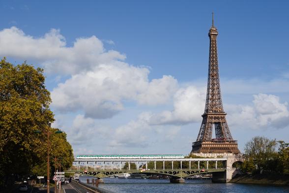 A view of the Eiffel Tower in Paris, from the Seine. On the left side of the picture, there is a large tree. The Bir Hakeim bridge, with a metro on it, appears prominently in the middle. On the right, the Eiffel tower immediately catches the eye. The sky is blue with clouds, and the water of the Seine river has a beautiful shade of blue.