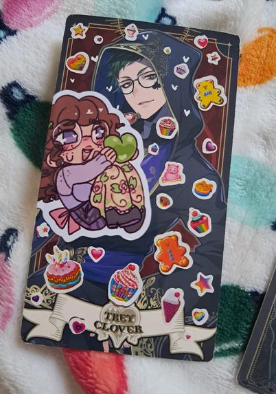 A card of Trey Clover in his ceremonial robes. It's decorated with a sticker of my OC Toli holding a heart toward his face as well as lots of hearts, stars, and desserts.