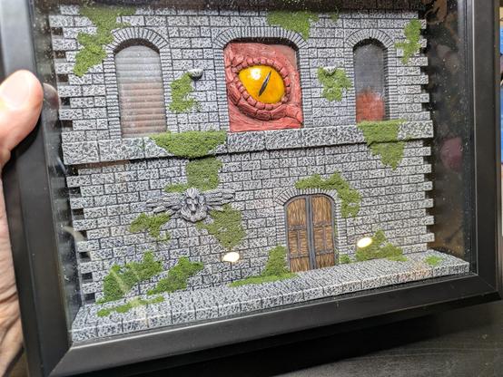 A diorama in a black box of a dungeon wall with moss now added. A dragon eye pokes out the top middle.