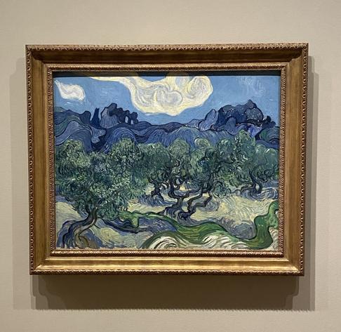 Ein gerahmtes Gemälde zeigt eine Landschaft mit Olivenbäumen und sanften Hügeln unter einem blauen Himmel mit wirbelnden Wolken. Das Kunstwerk zeichnet sich durch lebendige Farben und ausdrucksstarke Pinselstriche aus, die typisch für Vincent van Goghs Stil sind.