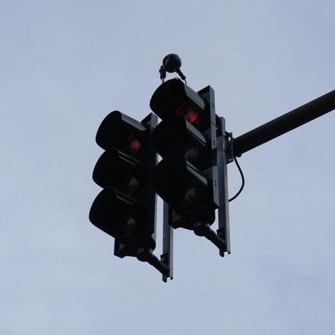The image shows a set of two traffic lights suspended by a metal pipe. The lights are on the red phase in the picture.
