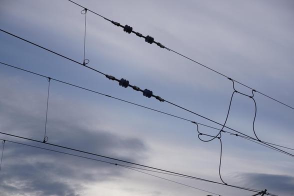 The image shows multiple train power cables with clouds in the background